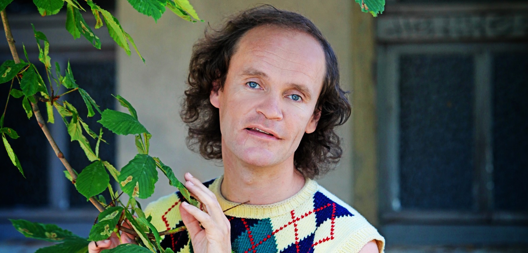 Olaf Schubert  in einem ärmellosen Pullover mit Argyle-Muster steht vor einem grünen Blätterhintergrund und hält vorsichtig einen Zweig. Er lächelt leicht und schaut direkt in die Kamera.