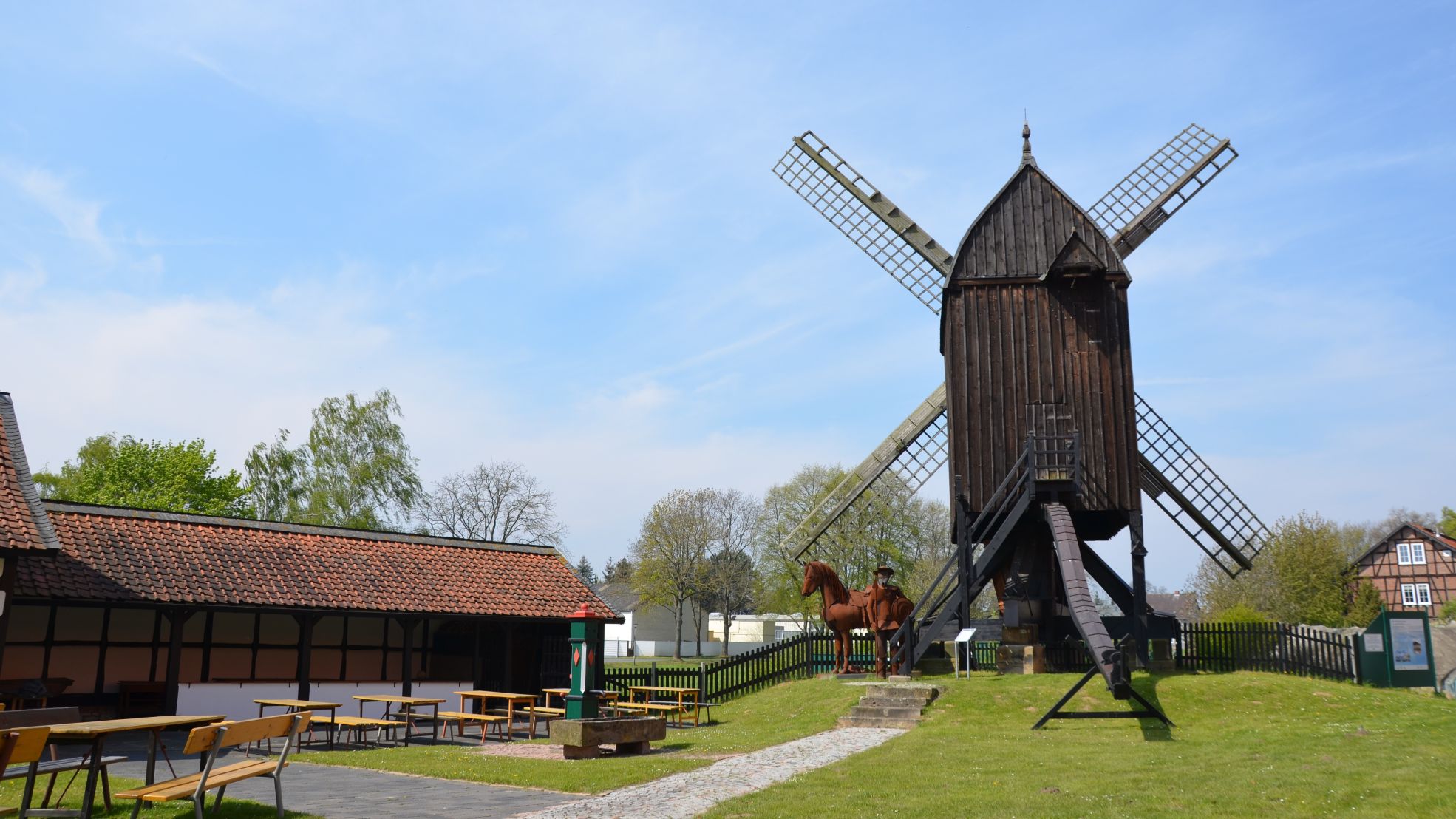 Historische Bockwindmühle mit Holzbau, flankiert von Sitzbänken und Fachwerkhaus im Grünen.