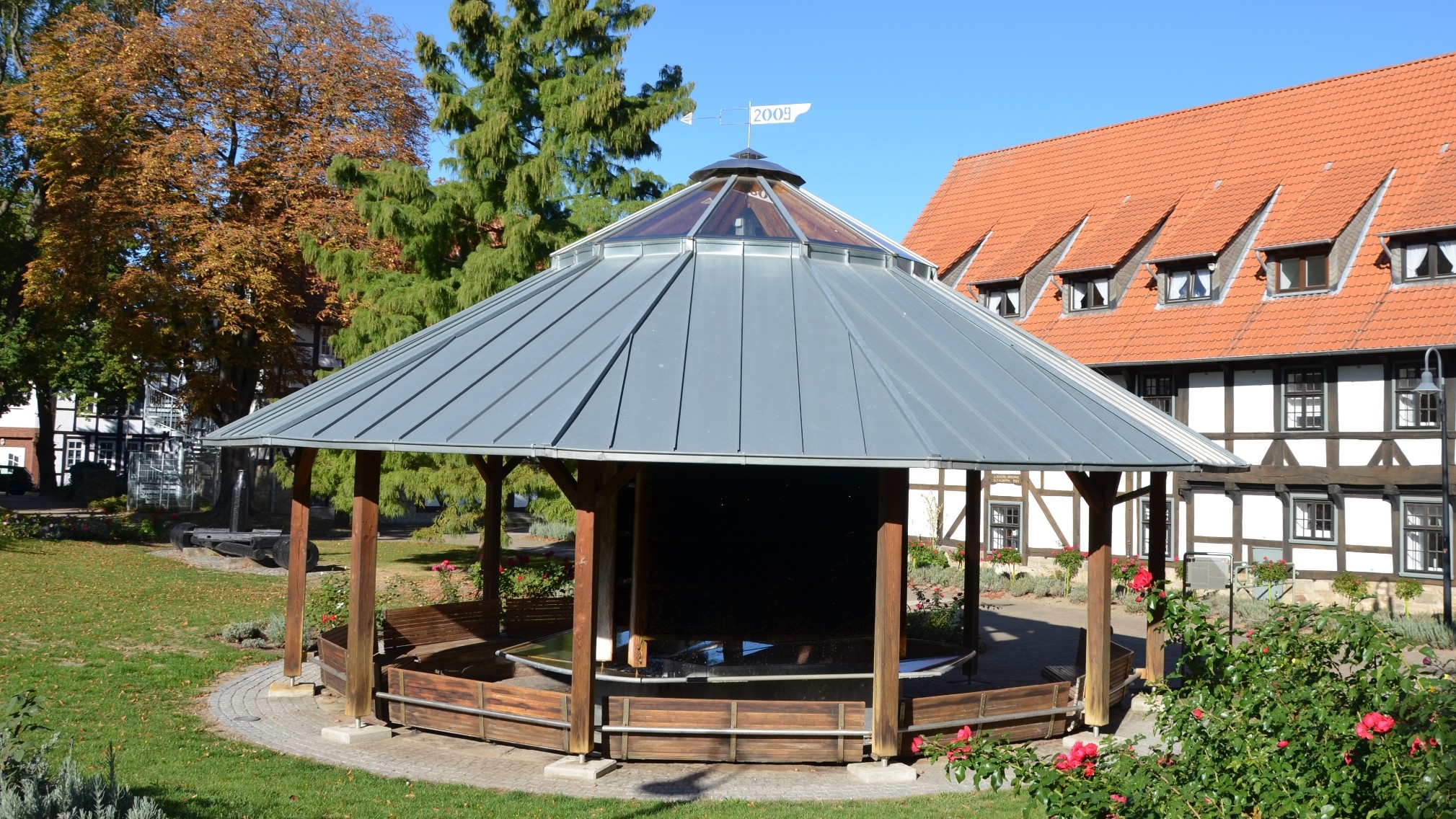 Runder Holz-Gradierpavillon mit Metalldach in einer Grünanlage vor einem Fachwerkhaus.